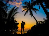Silueta de una pareja compartiendo un momento romántico en una playa tropical al atardecer con palmeras.