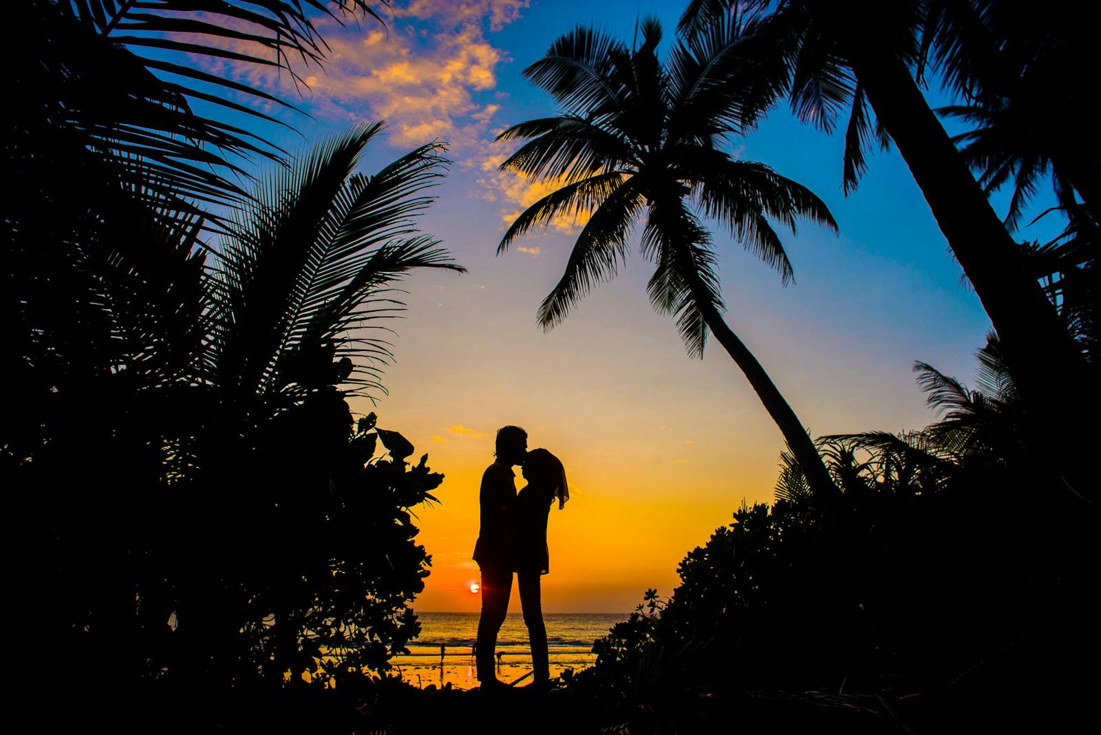 Silueta de una pareja compartiendo un momento romántico en una playa tropical al atardecer con palmeras.