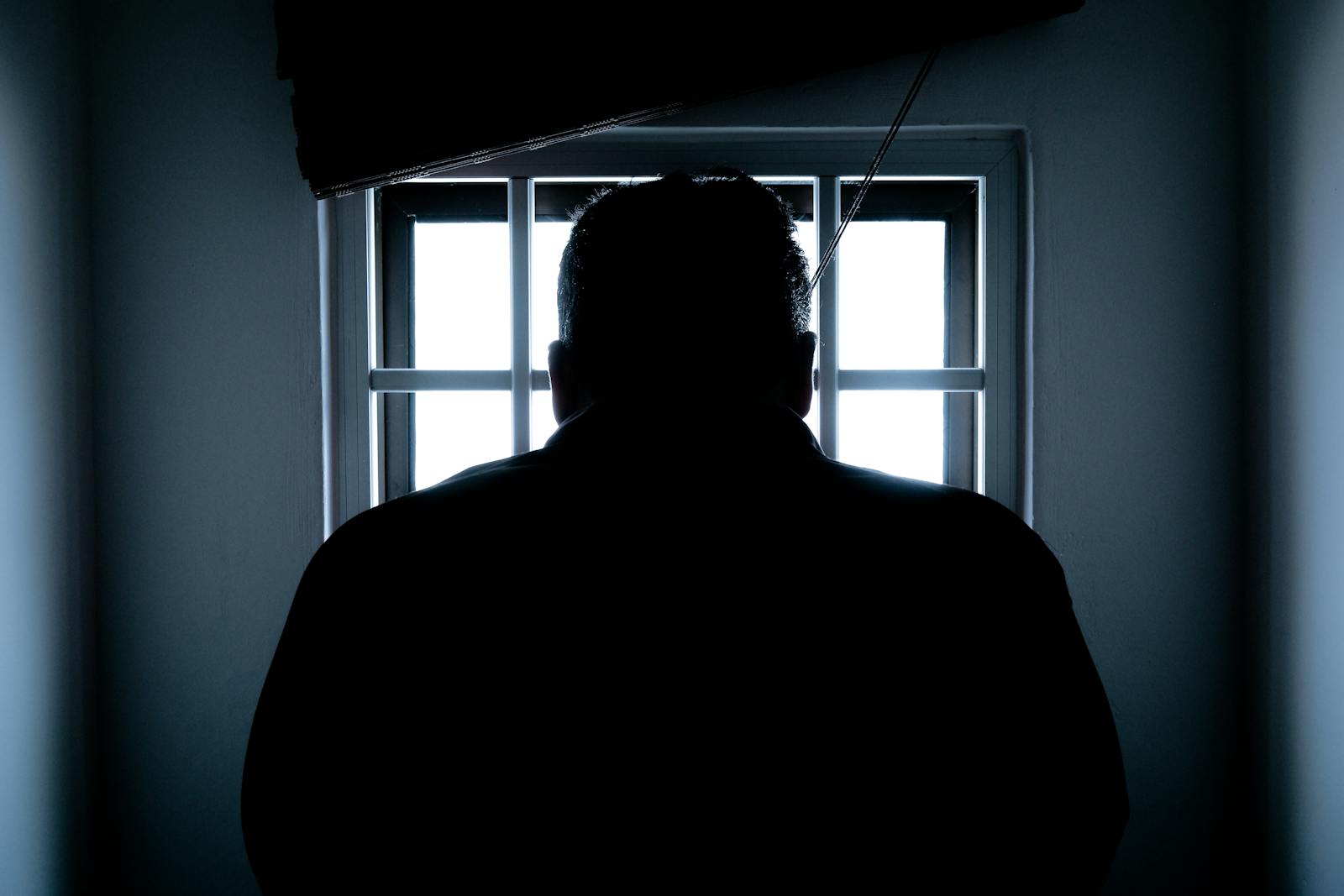 Silueta oscura de un hombre en una celda de prisión, iluminada desde atrás por los barrotes de la ventana.