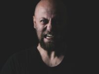 Primer plano de un hombre calvo y barbudo que muestra una expresión feroz en un entorno oscuro.